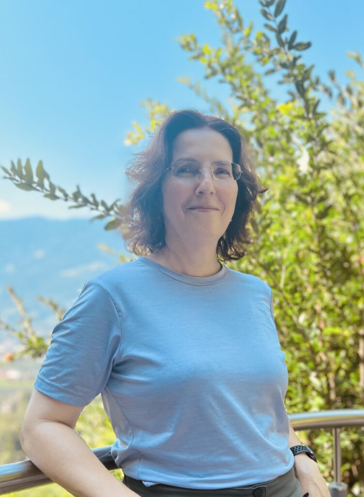 Woman in glasses and a light blue shirt stands by a railing with trees and mountains in the background.